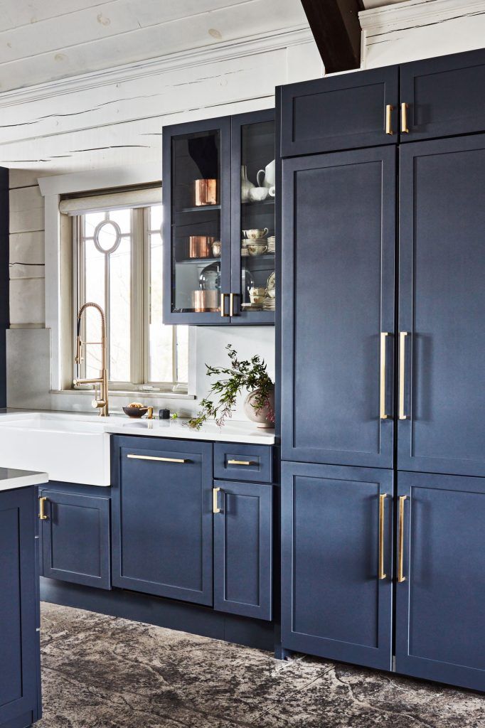 After the Remodeling: A modern kitchen with navy blue cabinets, brass handles, a farmhouse sink, and a brass faucet. A glass-front cabinet displays dishes; copper items sit on top. Light streams through a window above the sink, highlighting white walls and a rustic-style countertop.