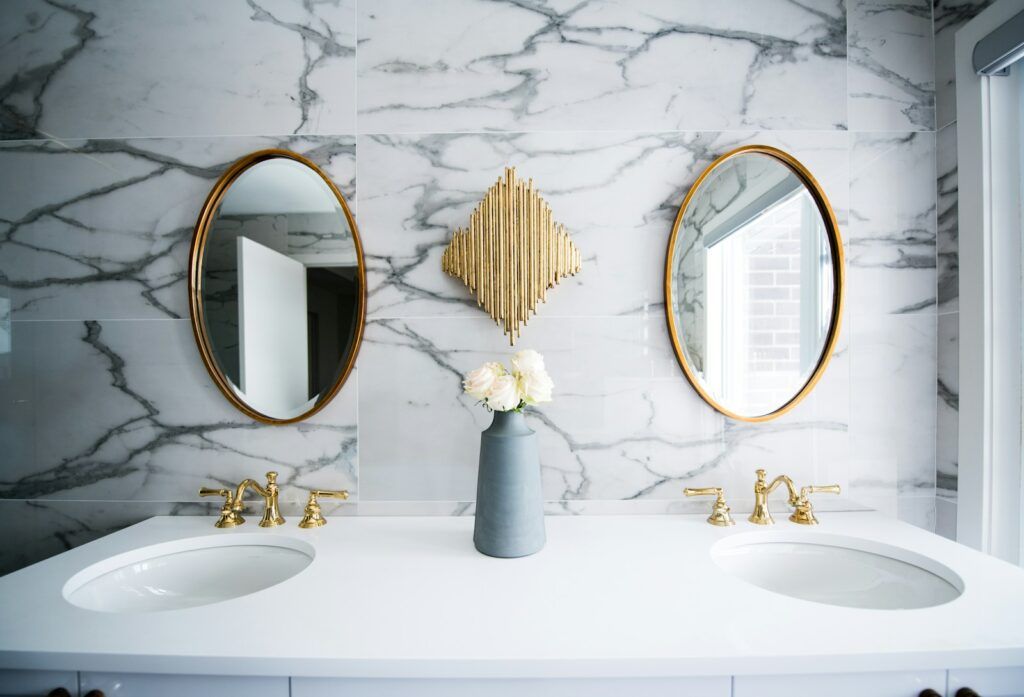 A modern bathroom by a skilled Bathroom Remodeler in Weehawken features a white marble wall, two oval gold-framed mirrors, gold faucets, and a double sink. A tall gray vase with white flowers sits beneath a gold wall piece as natural light streams in.