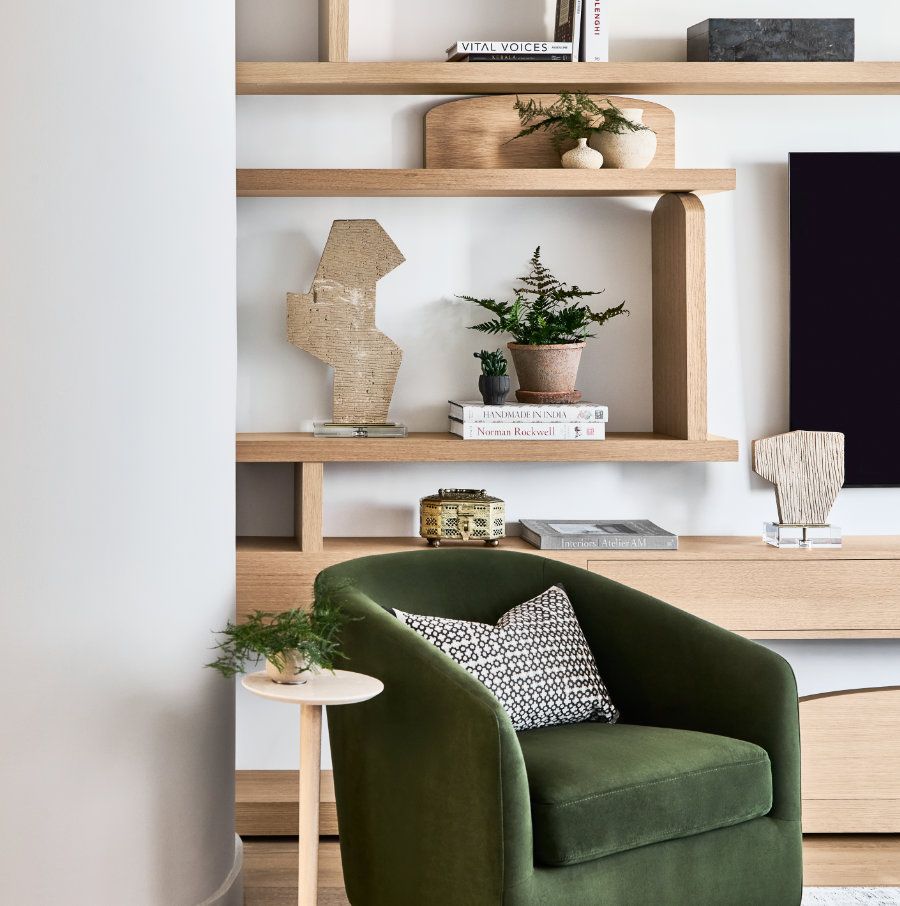 A modern living room corner, crafted by Avery Johnson Design, features open wooden shelves for books and décor, a green velvet armchair with a patterned pillow, and a round side table topped with a plant in a glass vase.