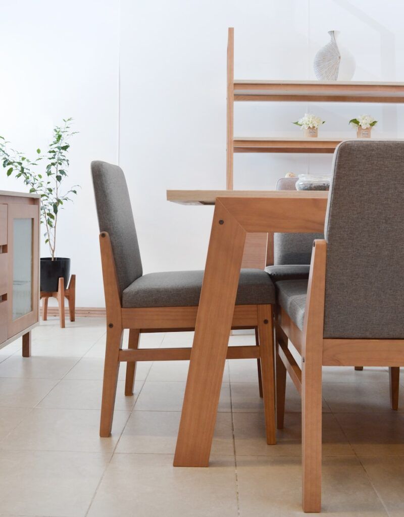 A modern dining area with light wood table and chairs featuring gray cushions. Showcasing thoughtful dining room design, a matching wooden shelf holds small plants and a ceramic vase. A potted plant stands on a light tiled floor against white walls.