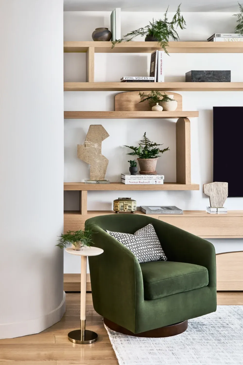 A modern living room corner designed by Avery Johnson Design, with a green velvet swivel chair, a small round side table with a gold base, and shelves behind holding plants, books, and decorative objects. Light wood flooring and neutral tones create a cozy, stylish atmosphere.