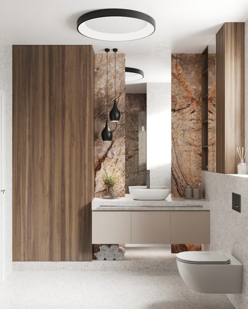 Modern bathroom by a Bathroom Remodeler in Weehawken featuring wood accents, a floating beige vanity, round white vessel sink, wall-mounted toilet, and black pendant lights. Marble wall with brown and gold veins adds striking contrast.