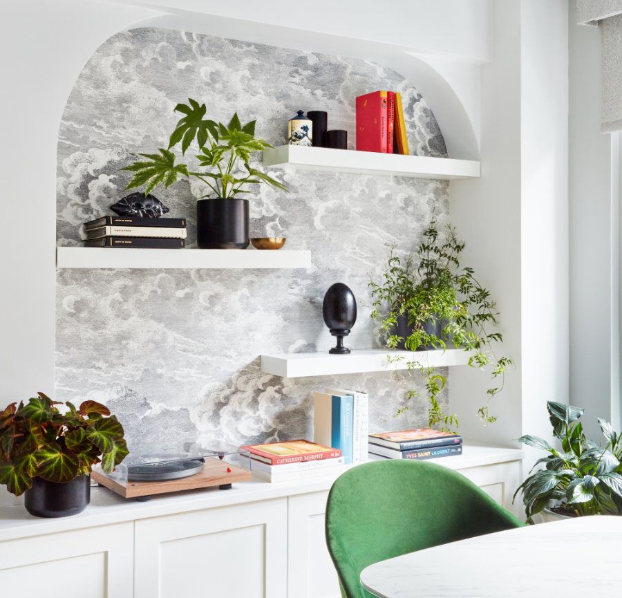 A modern nook, crafted by Avery Johnson Design, features two white floating shelves with books, black and white potted plants, decor accents, and a sculpture. Below, a built-in cabinet houses a turntable and books. A green velvet chair sits by a round table.