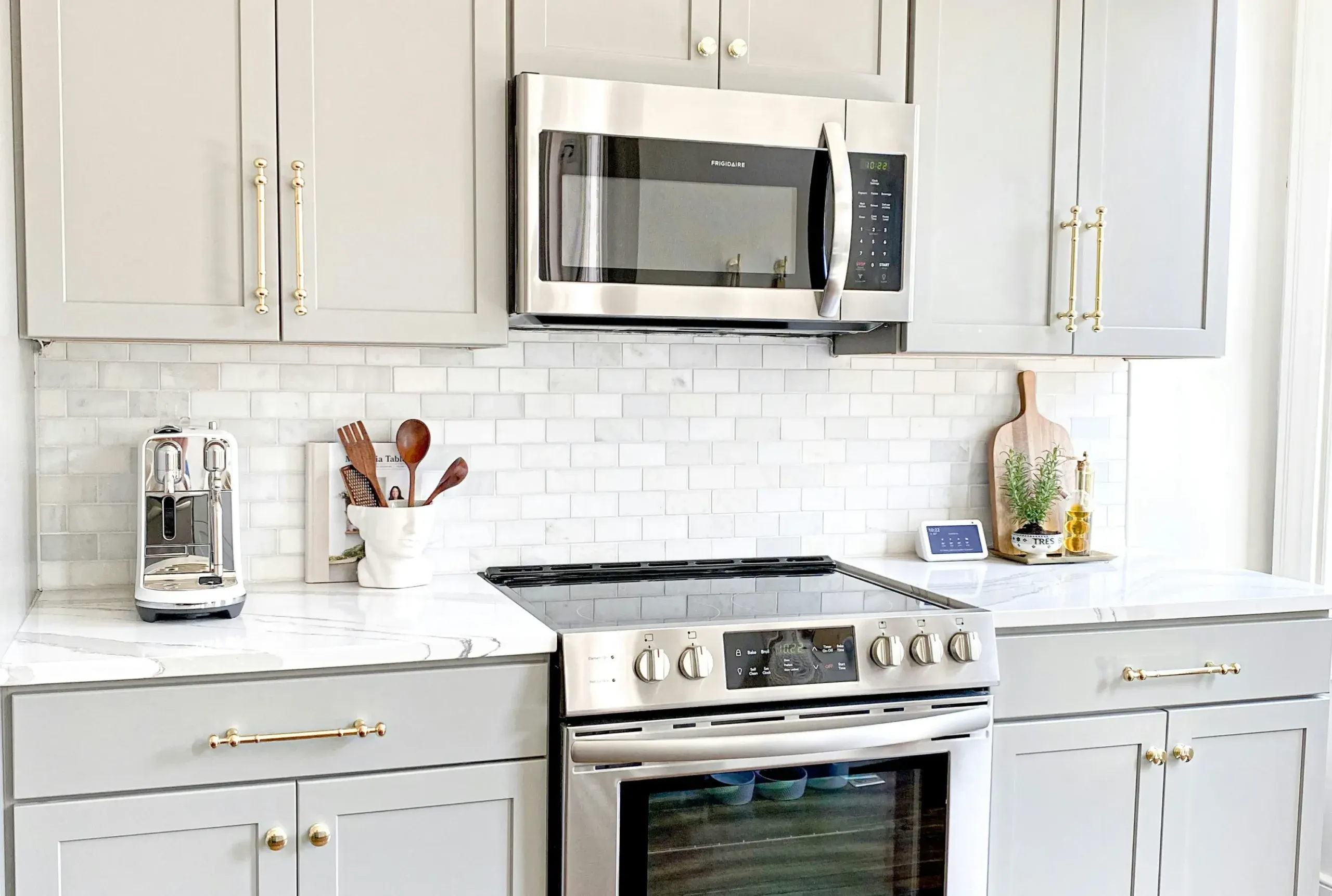Modern kitchen with light gray cabinets, stainless steel microwave above an electric stove, and white marble countertops. White subway tile backsplash, smart display, toaster, and fresh accents complete the look.