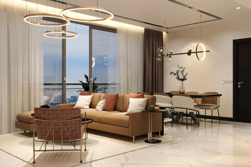 A modern living and dining area with beige and brown furniture, including a tan sofa, accent chair, and dining table with six white chairs. Circular pendant lights hang from the ceiling, and large windows are covered with sheer white curtains. Marble tile flooring.