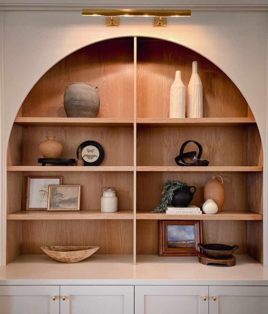 A built-in arched wood shelf with six compartments holds neutral-toned pottery, vases, framed art, books, a bowl, and decorative objects. The shelves are lit by a gold wall lamp above, highlighting elegant cabinetry & hardware design below.
