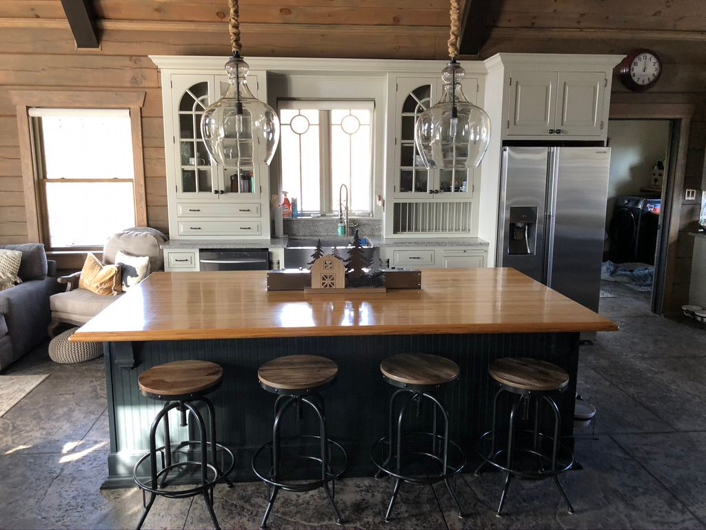 Before the remodeling: A rustic kitchen with a large wooden island, four round wooden stools with metal legs, hanging glass pendant lights, white cabinetry, and a farmhouse sink.