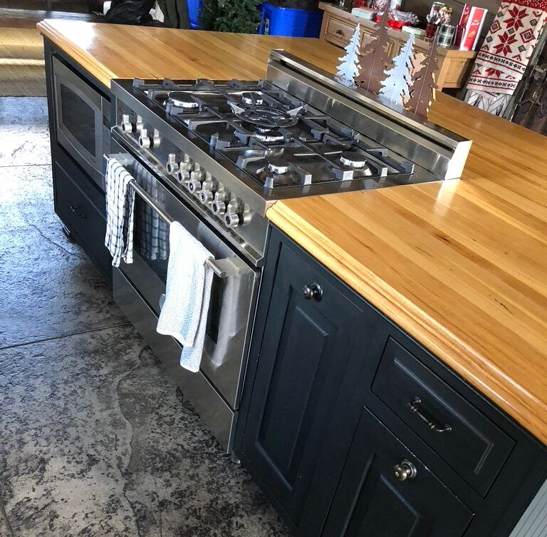Before the remodeling: A kitchen island with a wooden countertop features a stainless steel gas stove and oven, dark cabinetry, drawers, and a towel hanging from the oven handle.
