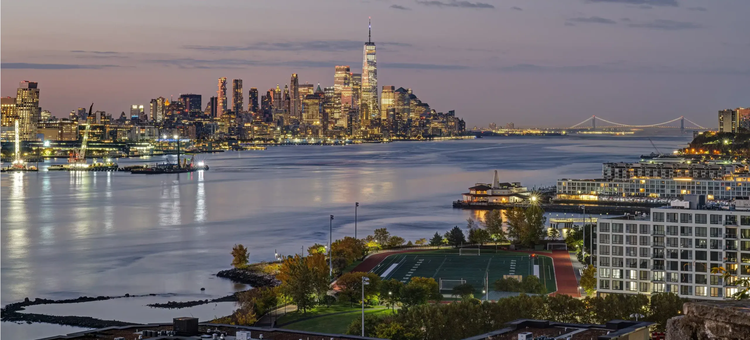 A sunset view of New York City’s skyline for Weehawken with illuminated skyscrapers reflected in the Hudson River, near a sports field and trees—just across from where a skilled Bathroom Remodeler in Weehawken transforms homes by the waterfront under a pastel sky.