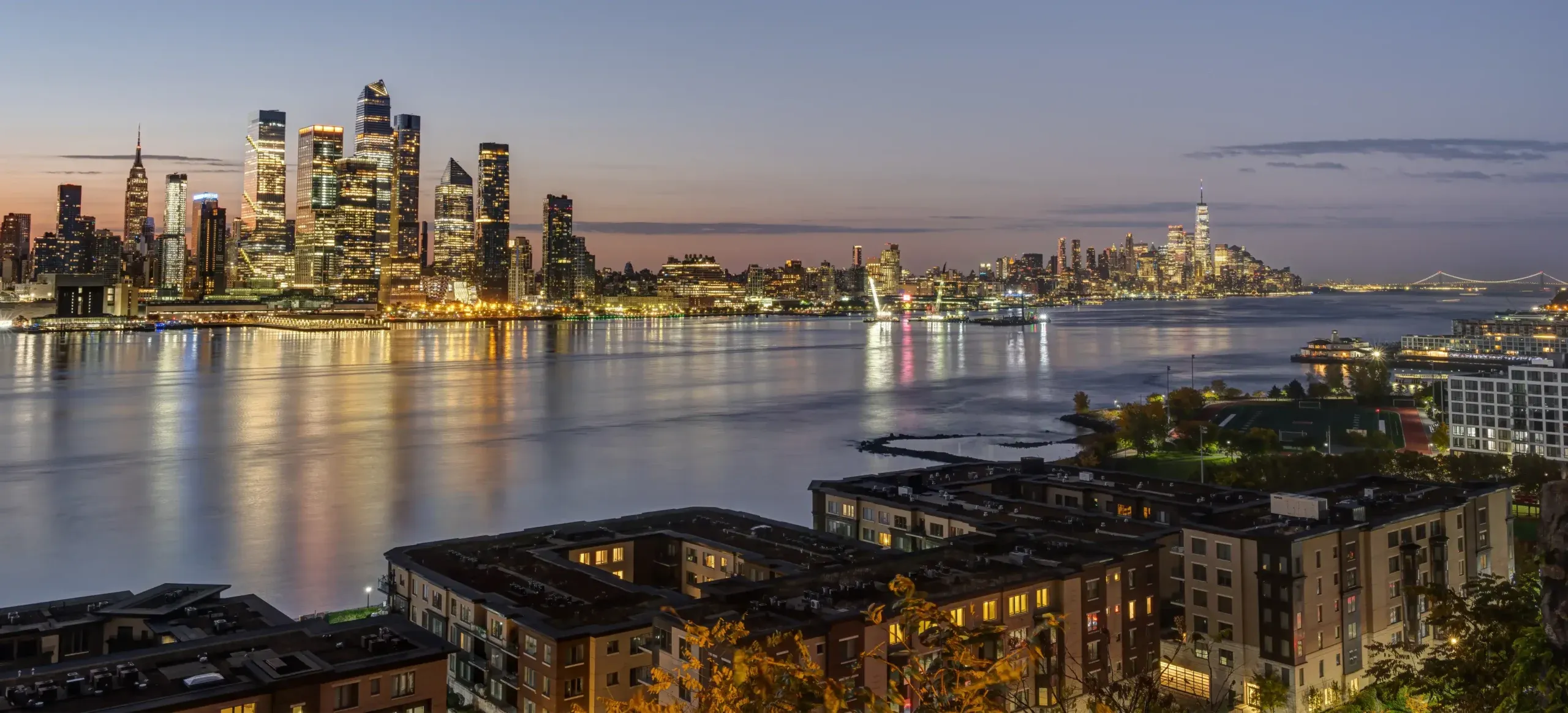 A twilight cityscape shows New York City’s skyline reflected in the Hudson River, with tall skyscrapers and glowing lights. In the foreground, rooftops and trees of residential buildings in Weehawken along the riverbank.