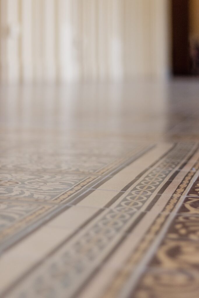 Close-up of ornate, patterned tile flooring in soft, neutral colors highlights an elegant flooring selection. The blurred background suggests a large, open indoor space with tall columns or drapes, creating a serene and refined atmosphere.
