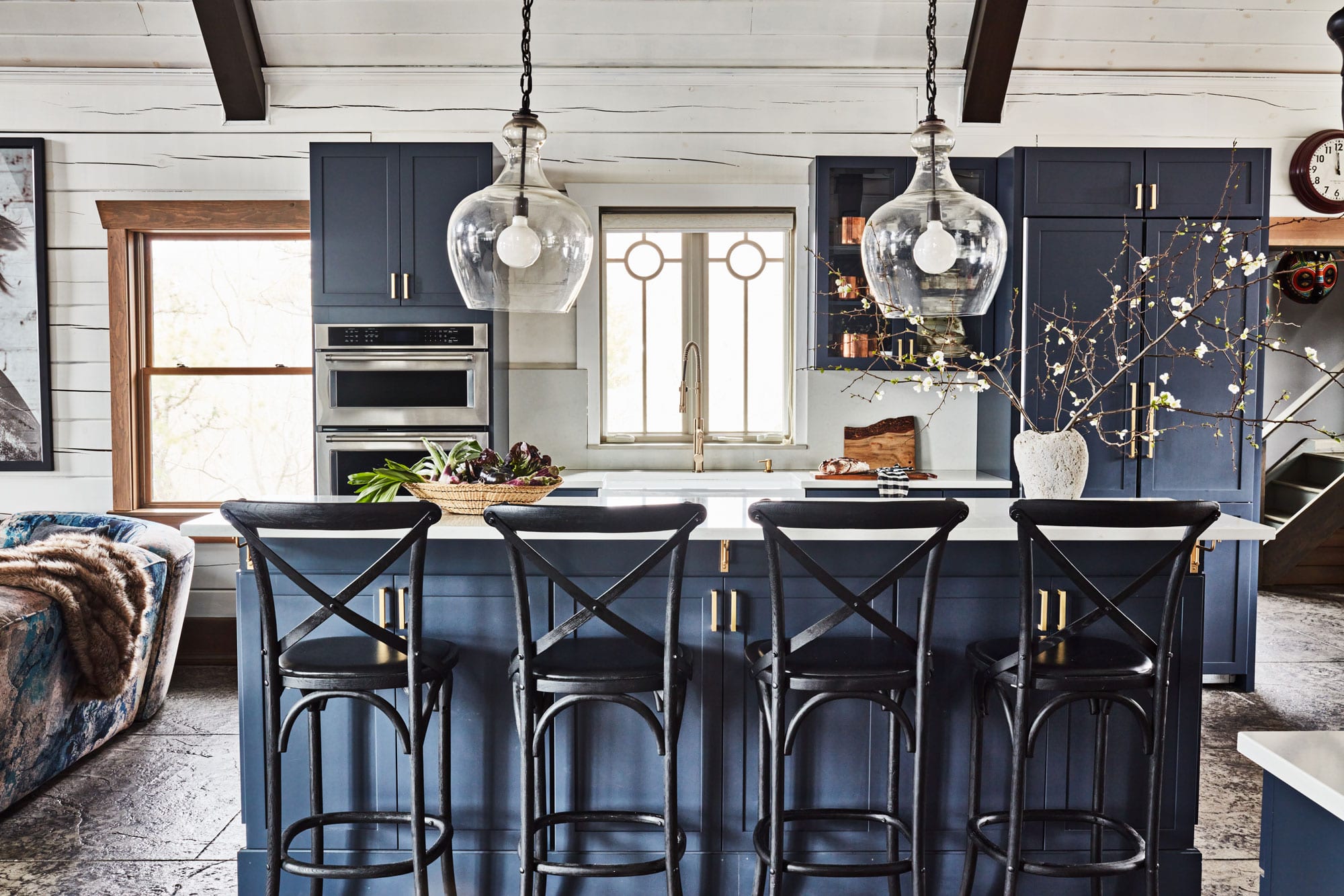 After the remodeling: A modern kitchen with blue cabinets, and a white island with four black high bar stools. Two glass pendant lights hang above the island. A vase with branches and a tray with plants sit on the counter. Sunlight streams through the window above the sink.